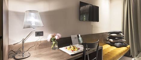 A hotel room with a bed, a desk with a lamp, a flat-screen TV, a vase with flowers, and a breakfast tray.