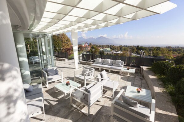 An outdoor patio with white furniture, glass walls, and a view of mountains and a cityscape.