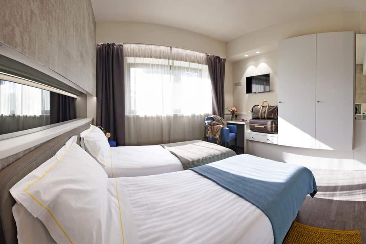 Habitación de hotel con dos camas, un escritorio, una silla, un armario y un amplio ventanal con cortinas.