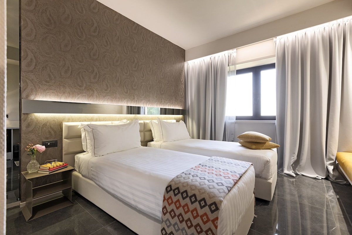 Una habitación de hotel moderna con dos camas, una mesita de noche con un florero y libros, y un ventanal con cortinas.
