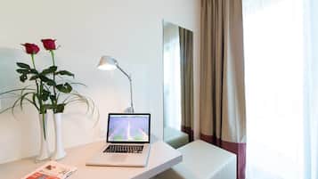 Ett modernt hotellrum med en bärbar dator på ett vitt skrivbord, en vas med två röda rosor, en golvlampa och ett stort fönster med gardiner.