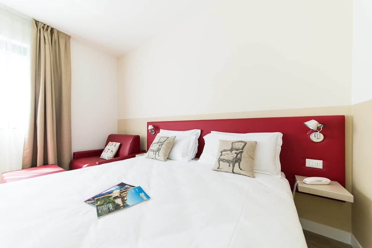 A hotel room with a large bed, a red headboard, white bedding, and a bedside table with a lamp and phone.