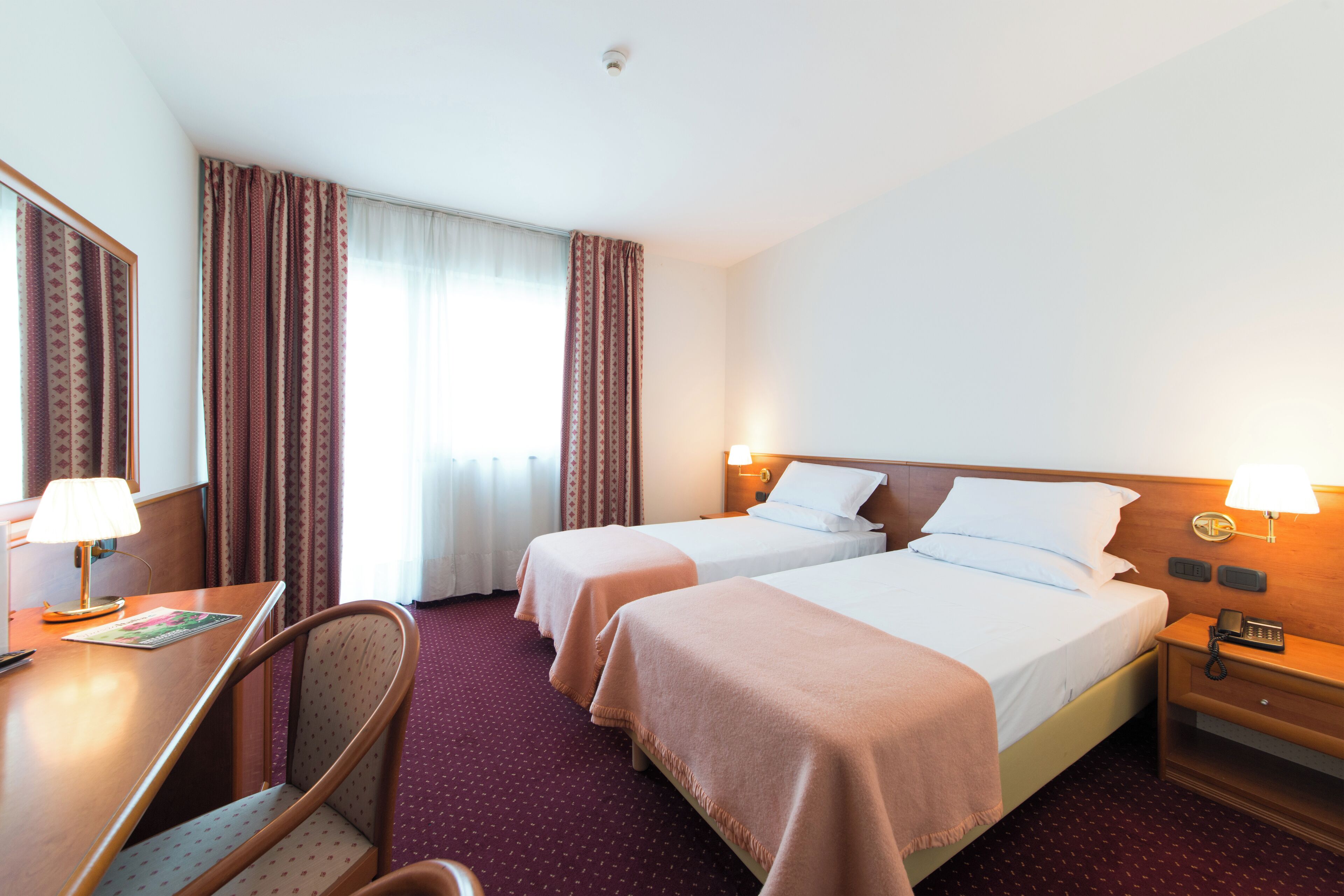 A hotel room with two beds, a desk, a chair, and a nightstand with a lamp.