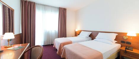 A hotel room with two beds, a desk, a chair, and a nightstand with a lamp.