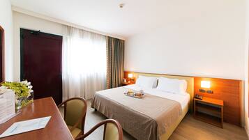 A hotel room with a bed, bedside table, lamp, and a desk with a chair.