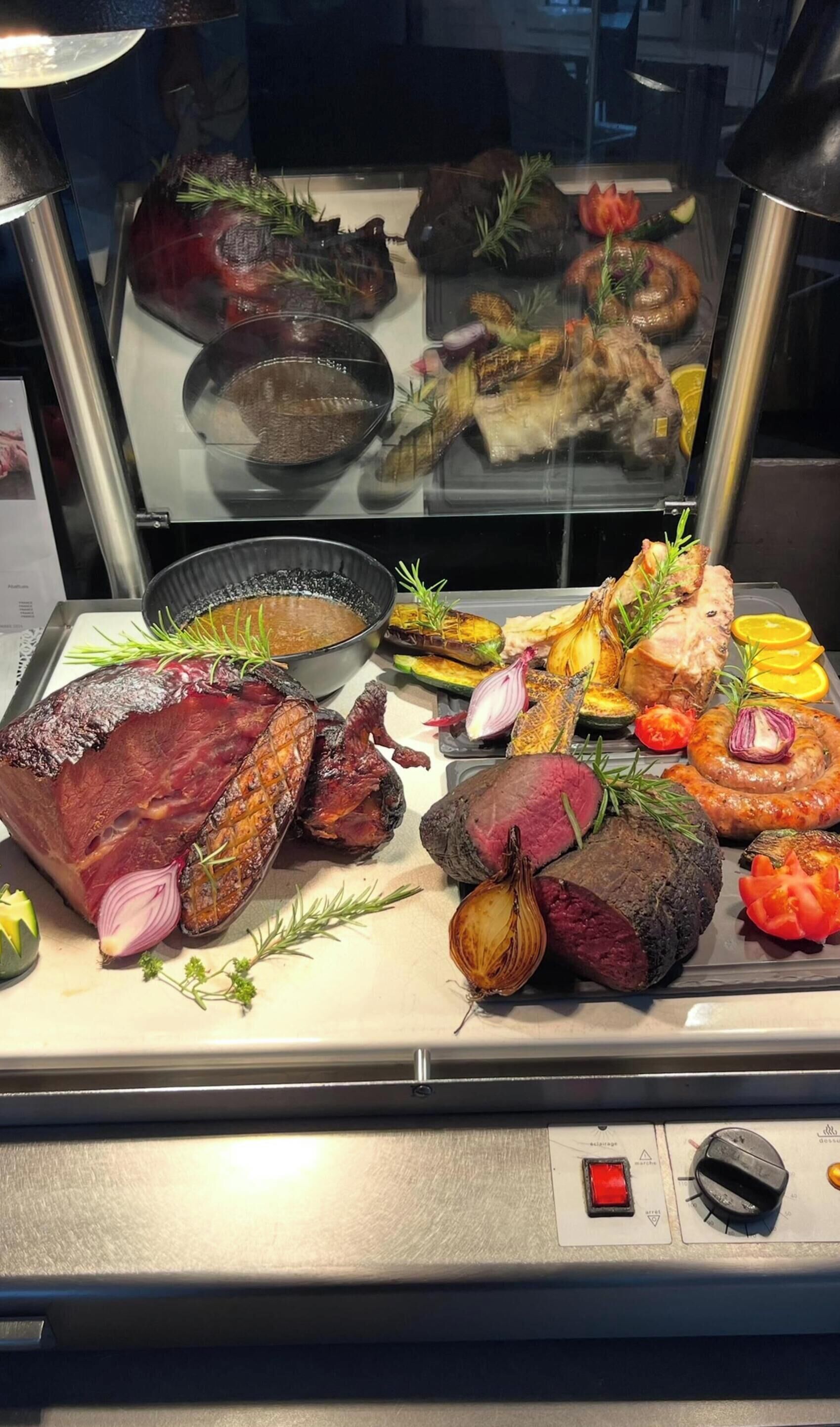 Vitrine com diversos tipos de carne e legumes, incluindo carnes assadas, salsichas e vegetais como cebolas e abobrinhas.