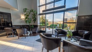 Un espace de détente intérieur moderne, avec des fauteuils en cuir et une table en verre, de grandes fenêtres offrant une vue sur l’extérieur, et des plantes en pot qui apportent une touche de verdure.