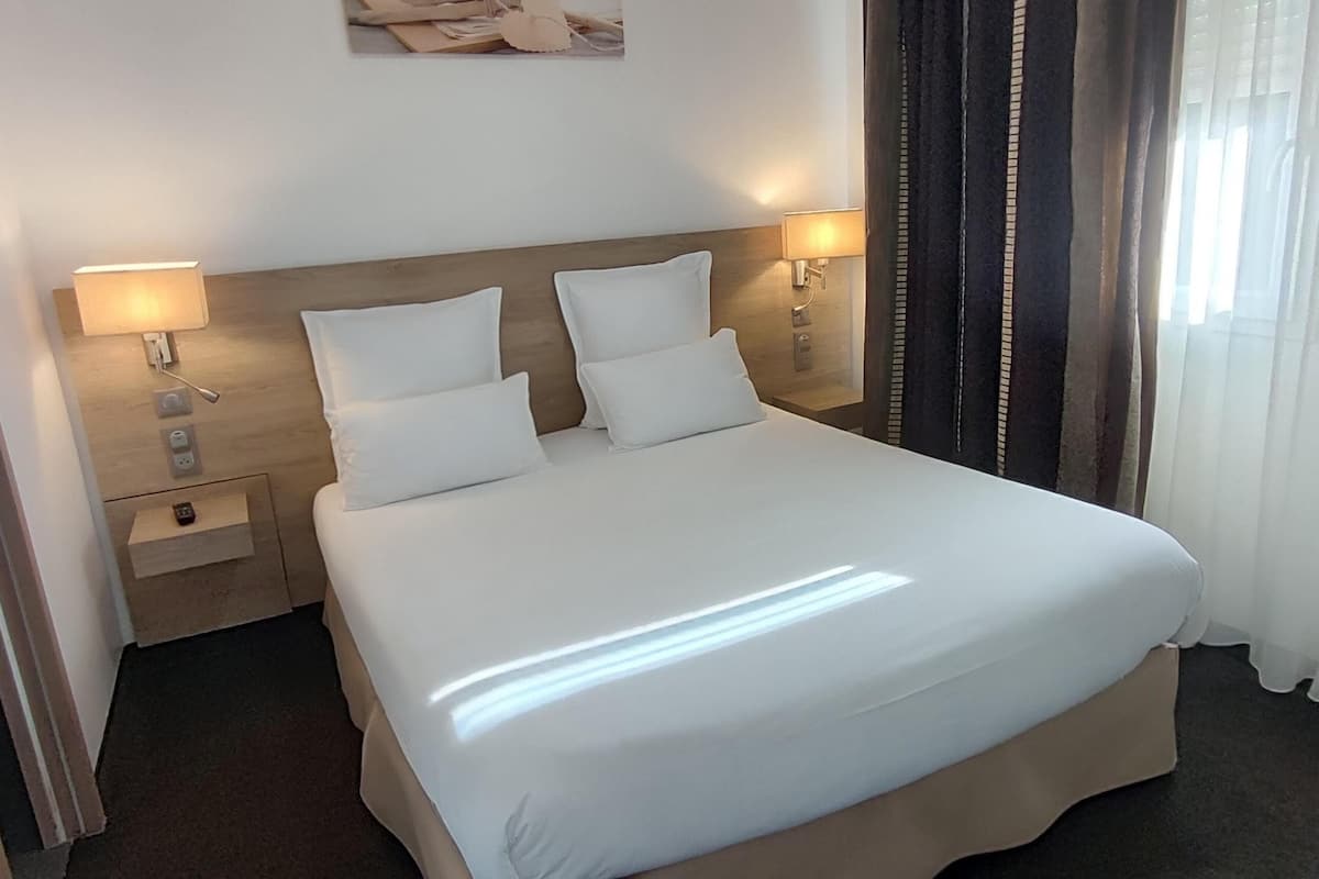 A neatly made bed with white linens, two bedside lamps, a nightstand with a phone and a small drawer, and a window with sheer curtains.