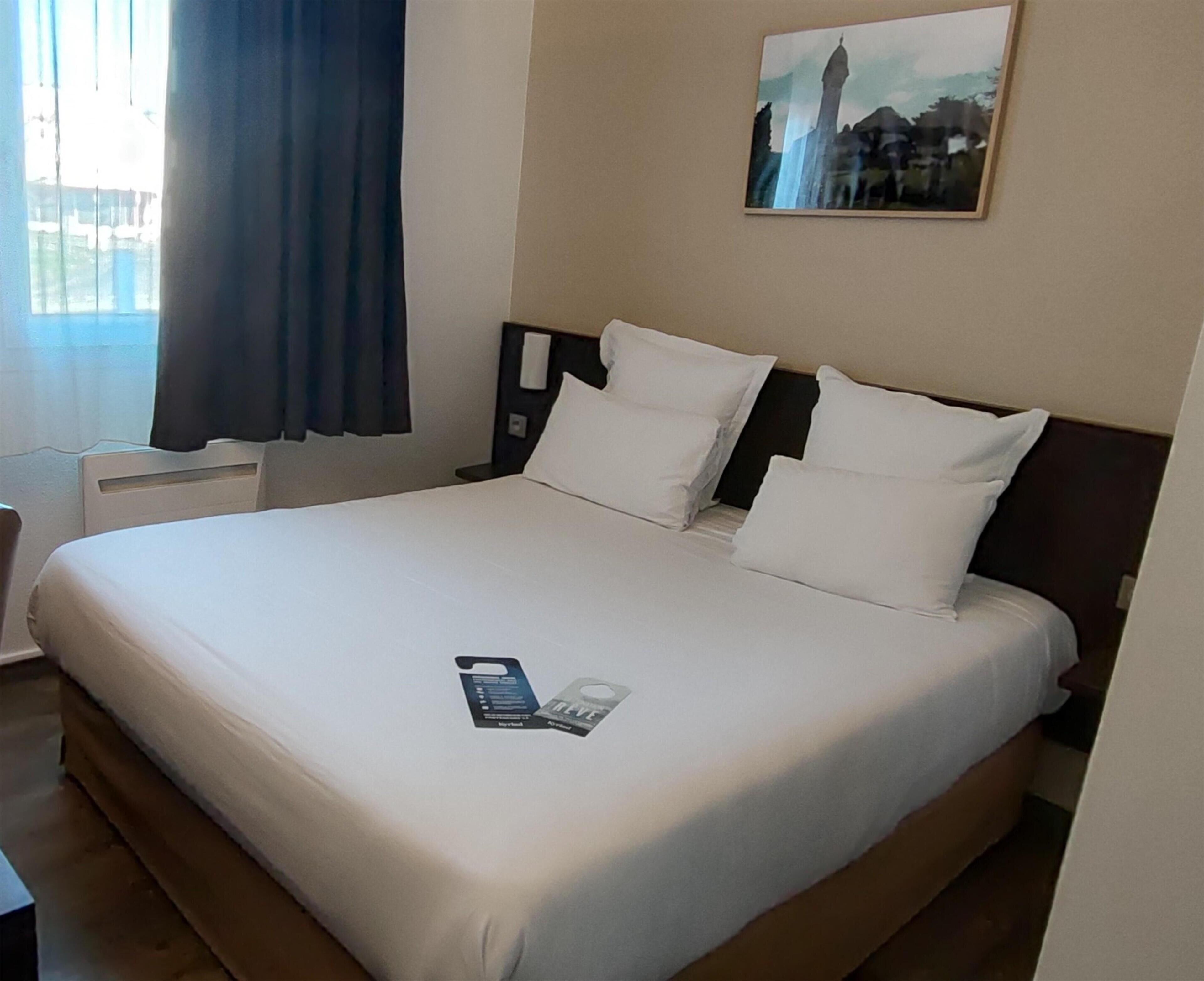A hotel room with a large bed, a nightstand, a wall-mounted phone, and a window with curtains.