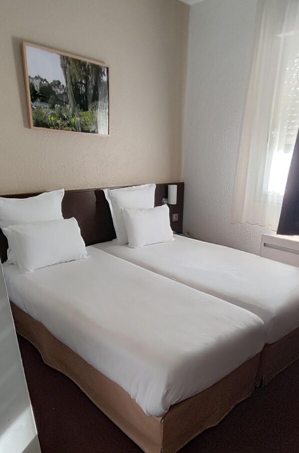 Una cama doble con sábanas blancas, cabecera de madera, un cuadro en la pared y una ventana con cortinas.