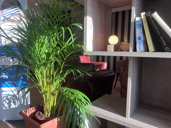 Une plante en pot, une bibliothèque remplie de livres, une lampe et un fauteuil rouge dans une pièce dont le mur est rayé et le plancher en bois.