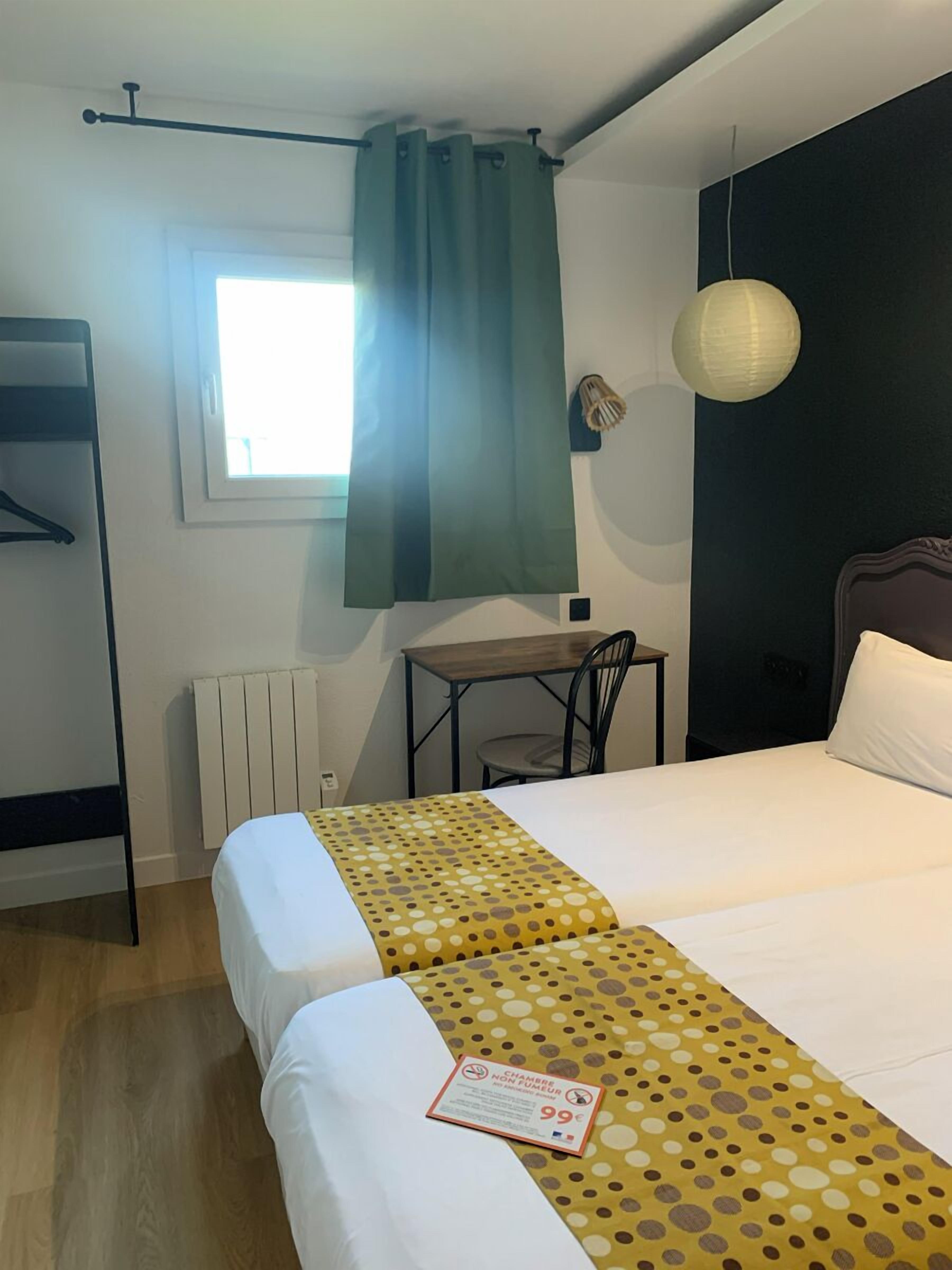 A hotel room with a bed, a desk, a chair, a radiator, a window with curtains, and a hanging light fixture.