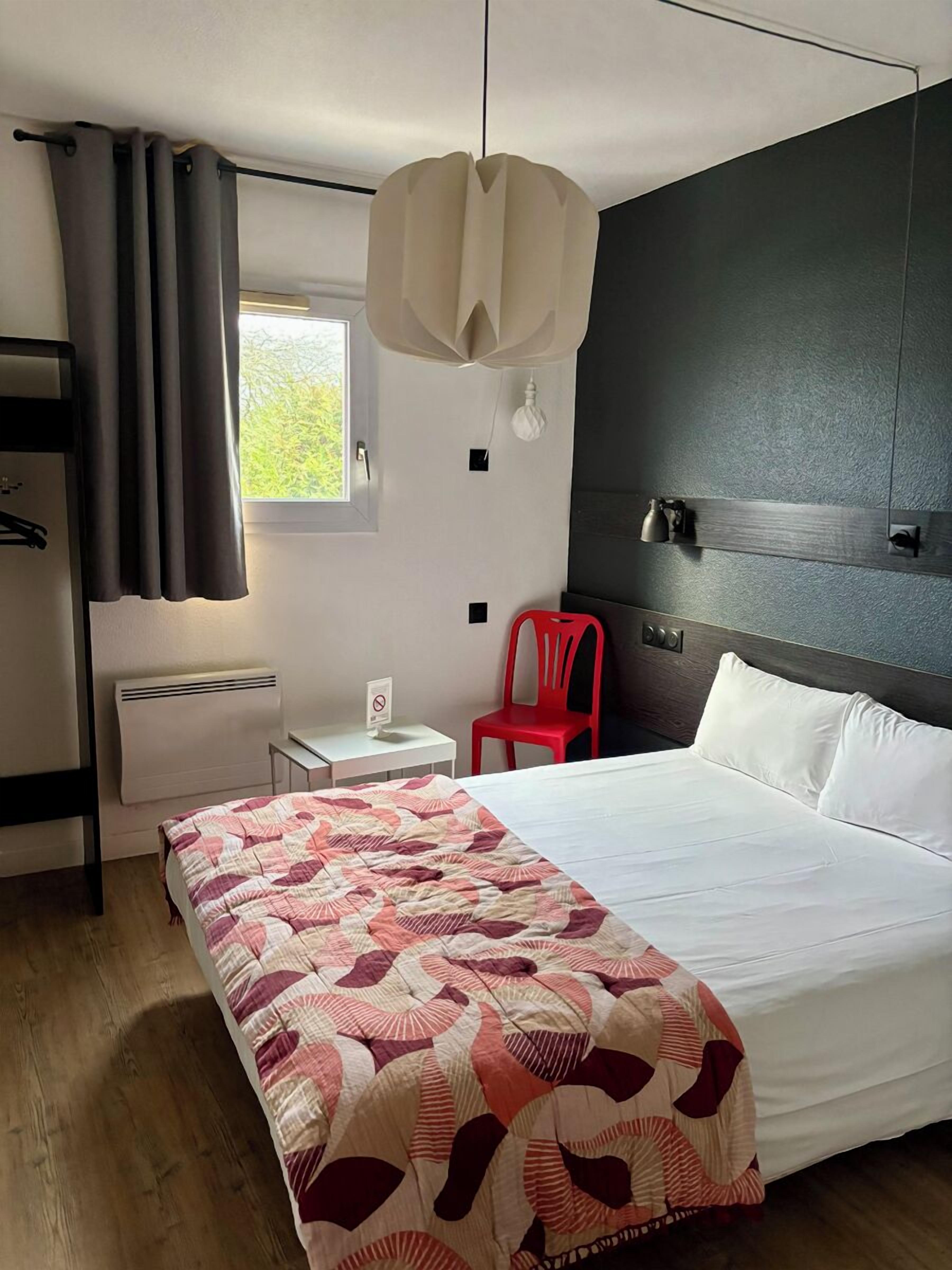 Une chambre à coucher comprenant un lit, un bureau, une chaise, une fenêtre et un luminaire fixé au mur.