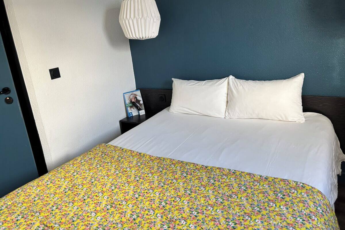 Un lit avec un drap blanc et une couverture à motifs floraux colorés, une lampe suspendue blanche et un mur bleu foncé.
