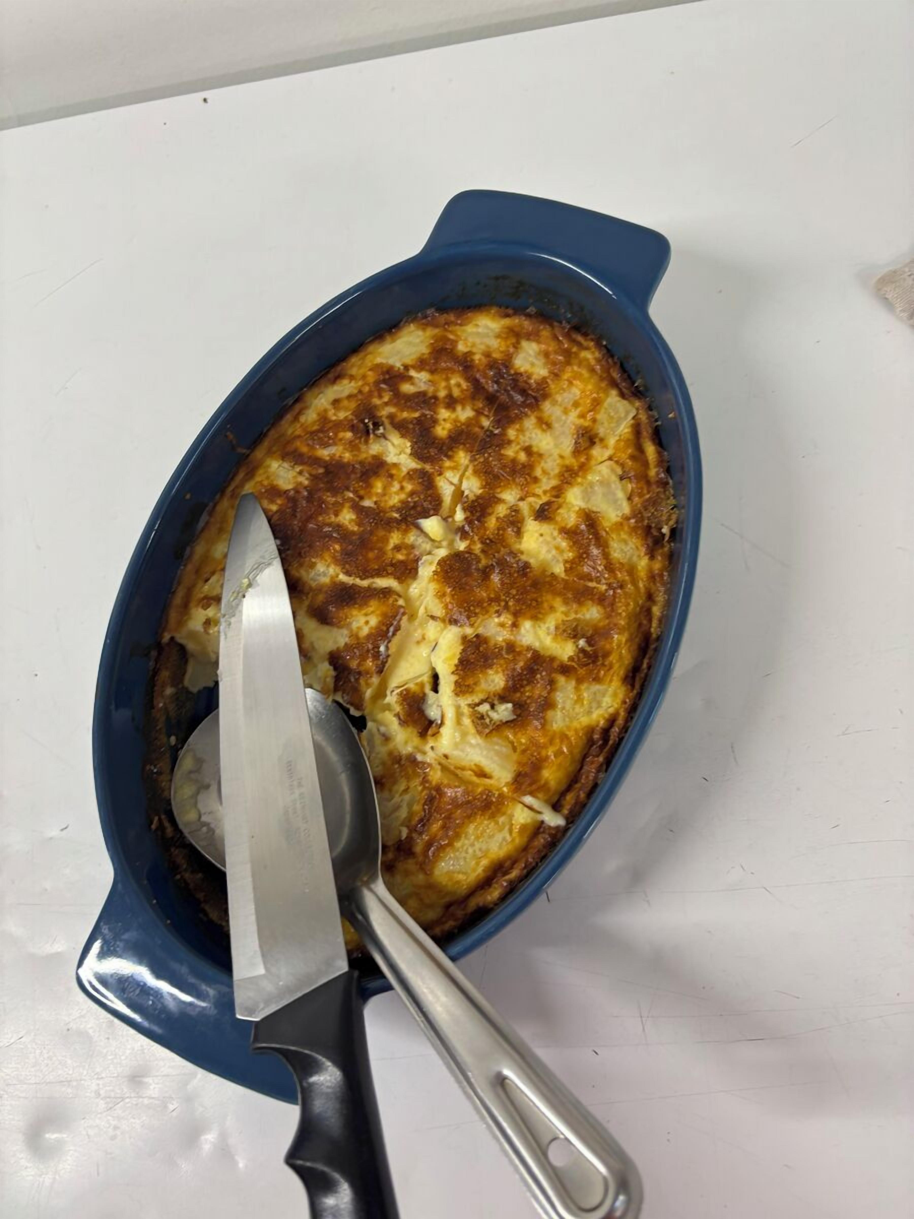 A blue baking dish with a golden-brown baked dish inside, a knife, and a spoon on a white surface.