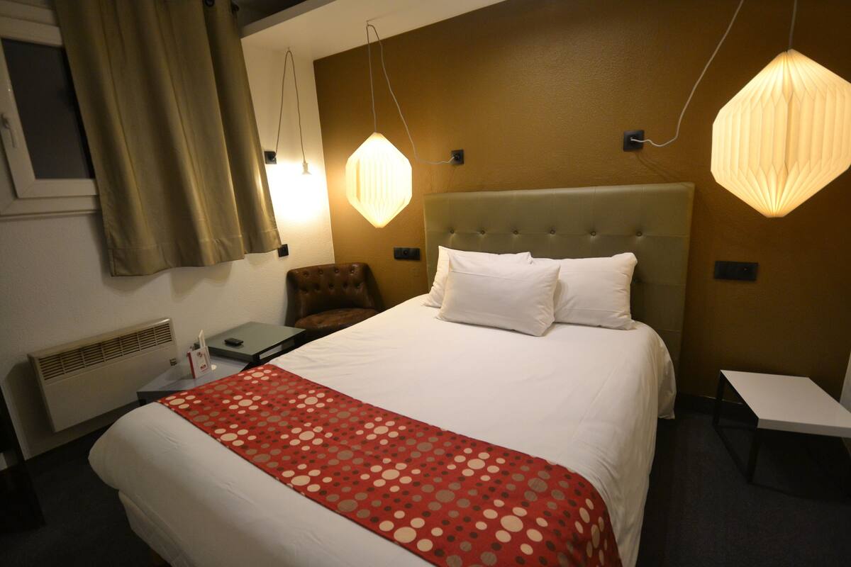 Une chambre d’hôtel avec un lit, une chaise, un bureau et deux lampes suspendues.