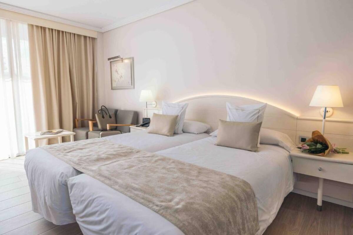 A hotel room with two beds, a desk, a chair, a lamp, and a bedside table with a bouquet of flowers.