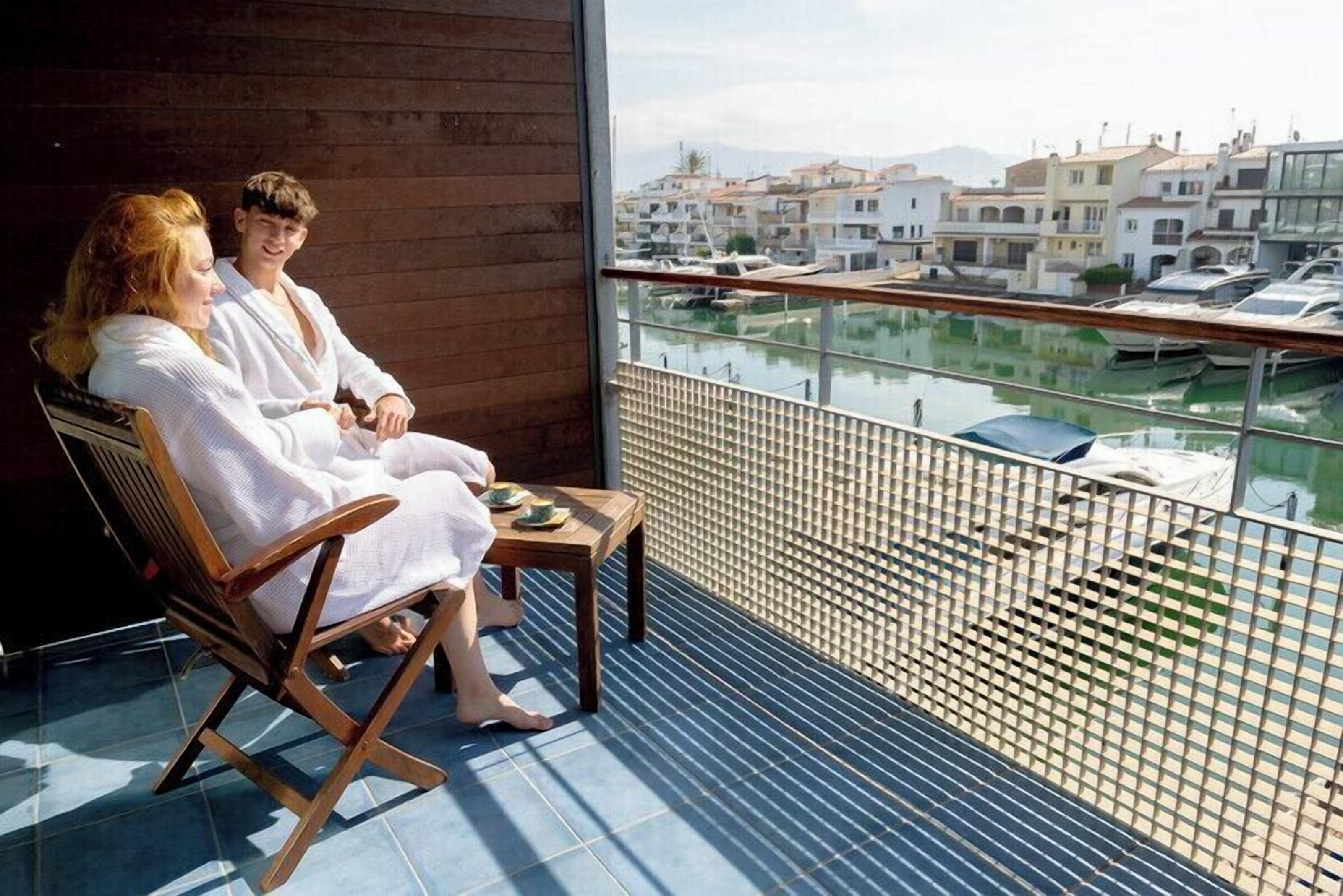 Un balcon donnant sur les bateaux et les bâtiments, avec des chaises en bois et une table où sont posées des assiettes.