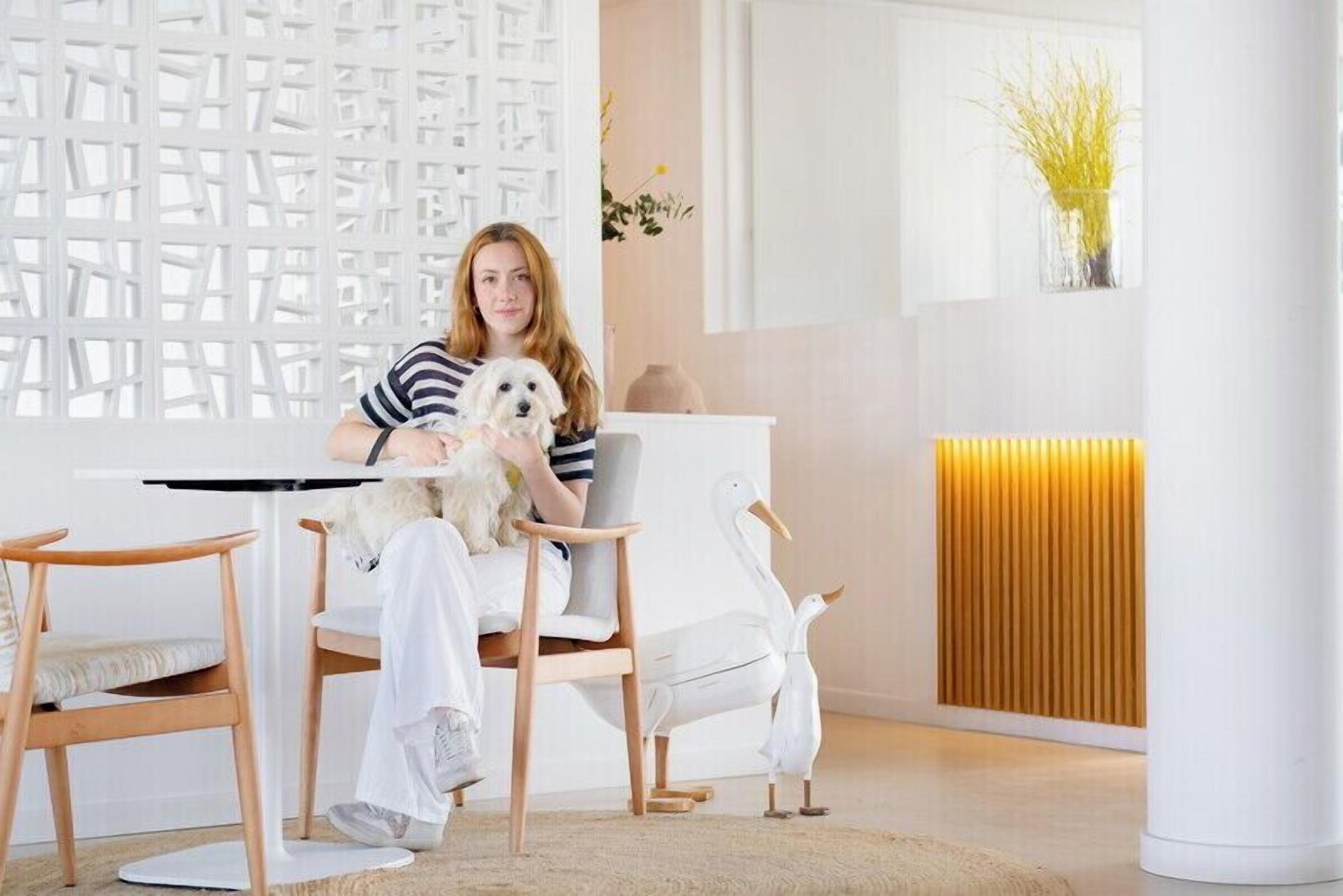 Een persoon zit aan een witte tafel, met een kleine hond, omringd door houten stoelen en een witte stoel, in een kamer met een witte muur en een houten paneel.