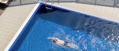 A swimming pool with a swimmer, deck chairs, and a tiled deck.