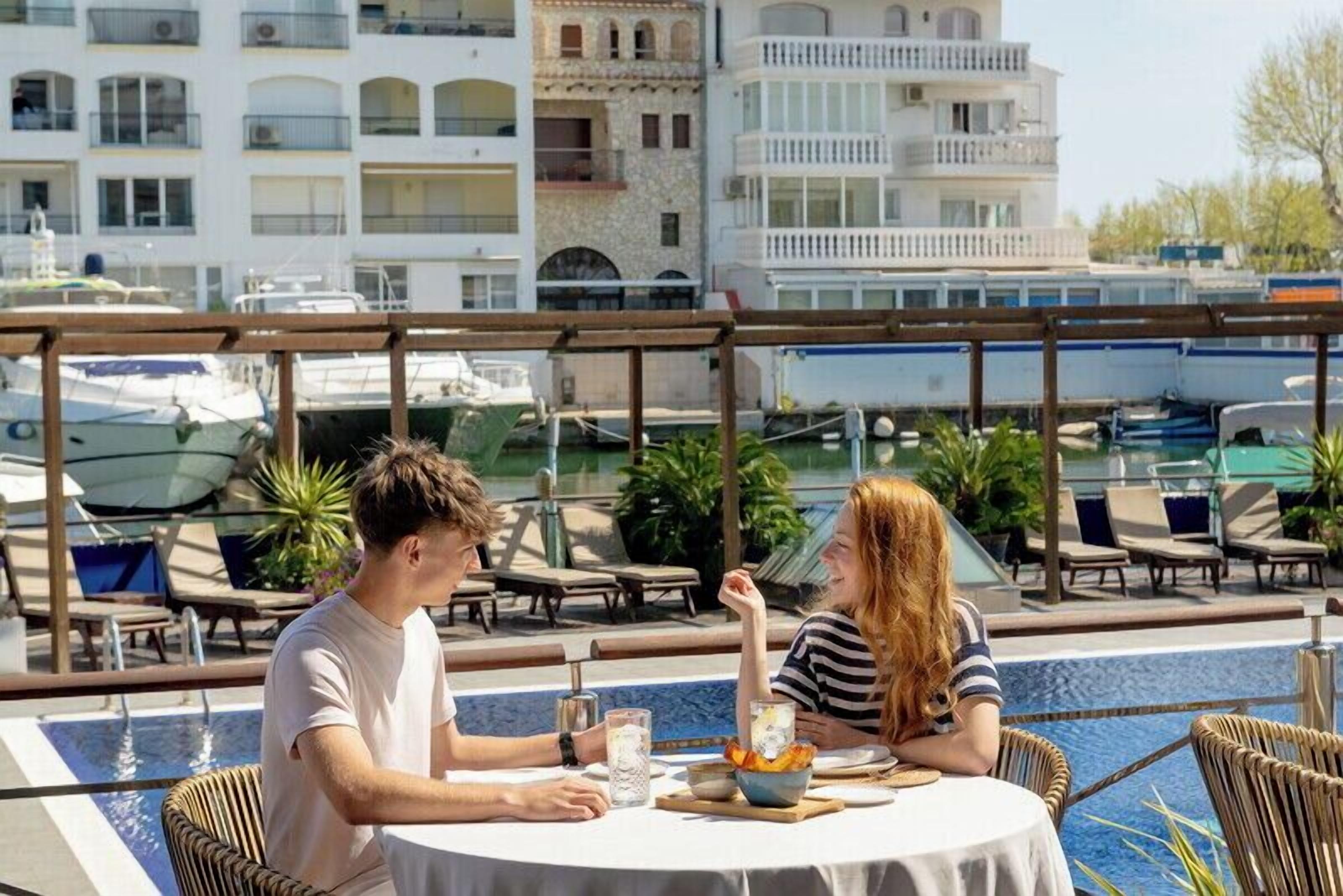 Una pareja cenando en una mesa redonda junto a una piscina, con vistas a barcos y edificios al fondo.