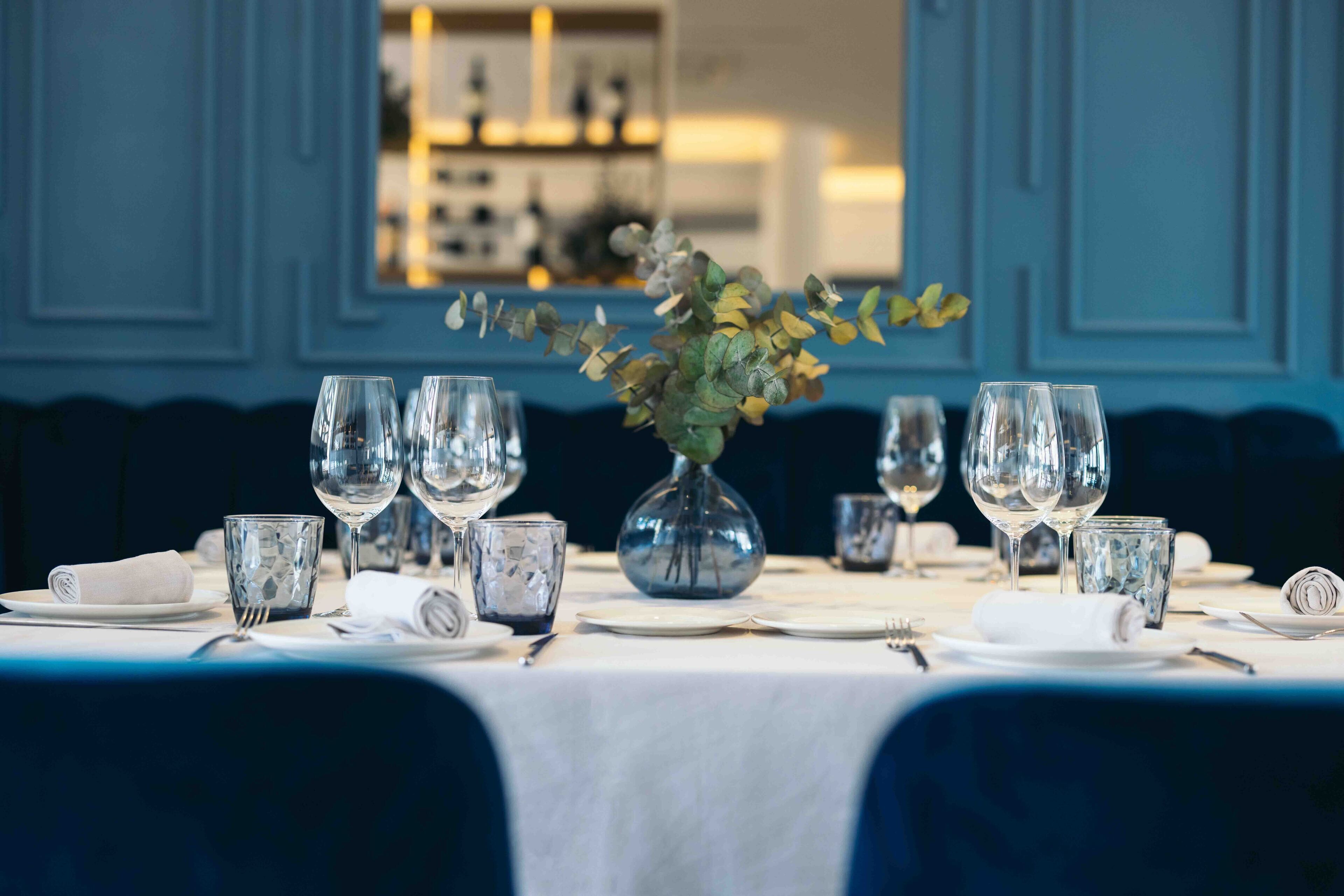 Una mesa de comedor dispuesta con varias copas de vino, un florero con follaje y servilletas dobladas.