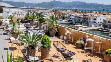 A rooftop terrace with lounge chairs, potted plants, and a view of the sea and buildings.