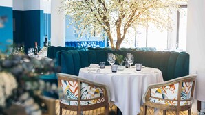 A dining area with a round table set for two, upholstered booths, and a large tree with white blossoms in the background. - Hotel Port Salins by Pierre & Vacances (Castello d'Empuries)