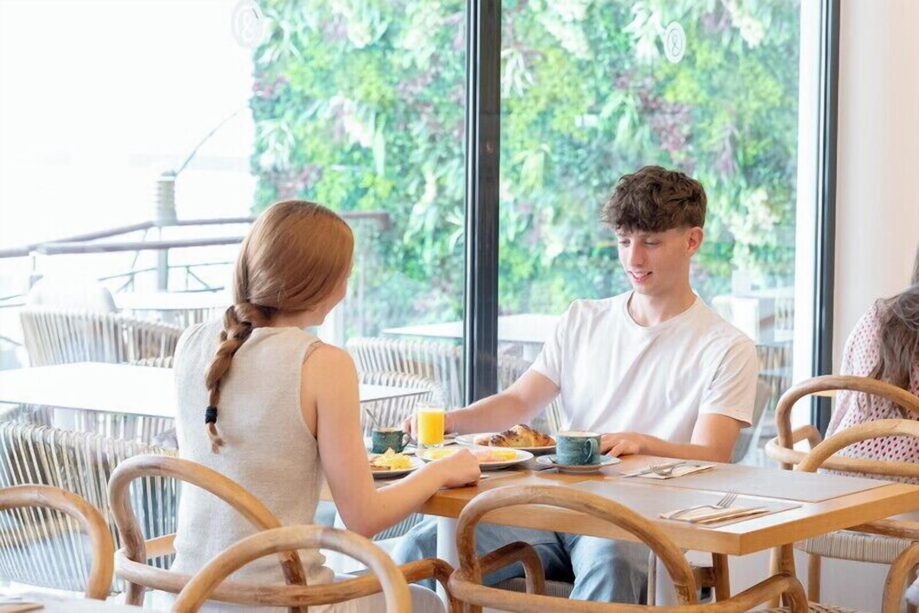 A breakfast setting with a table, chairs, and a view of greenery through large windows.