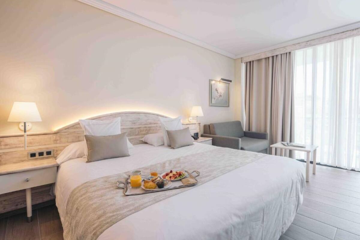 A hotel room with a large bed, bedside lamps, a small table with breakfast, and a view of the outdoors through large windows.