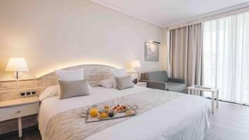 Habitación de hotel con una cama grande, lámparas de noche, una pequeña mesa con desayuno y vistas al exterior a través de amplios ventanales.