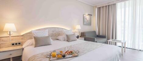 Habitación de hotel con una cama grande, lámparas de noche, una pequeña mesa con desayuno y vistas al exterior a través de amplios ventanales.