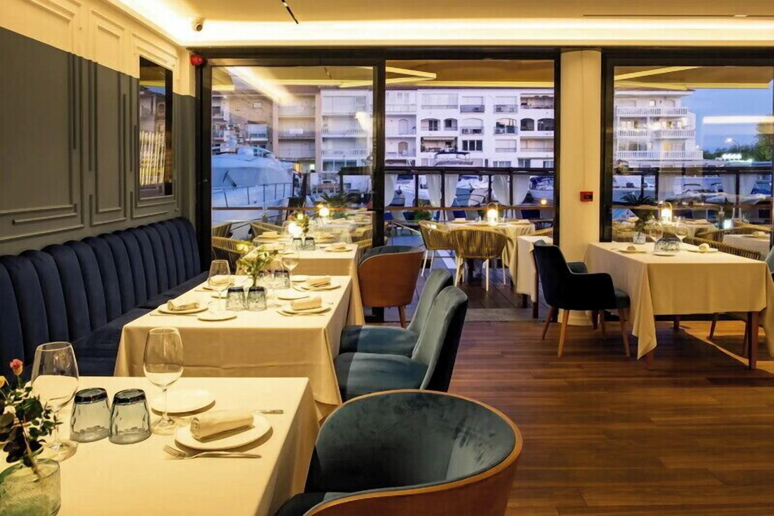 En restaurant med dækkede borde, et siddeområde med bænk og udsigt til bygninger gennem store vinduer.
