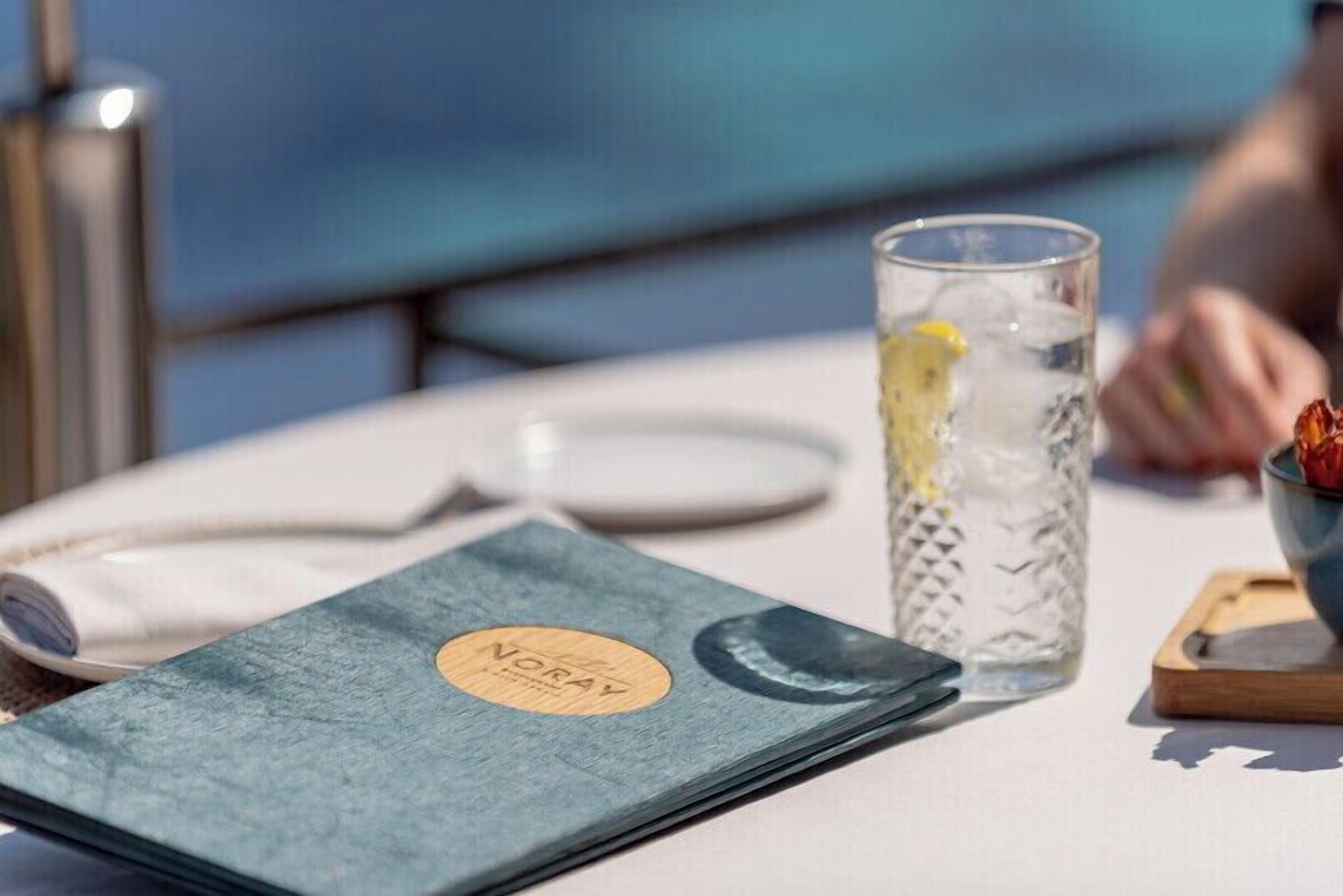 A table with a menu, a glass of water with a lemon, and a bowl of fruit.