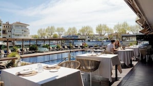 An outdoor dining area with tables and chairs overlooking a swimming pool, with buildings and trees in the background. - Hotel Port Salins by Pierre & Vacances (Castello d'Empuries)