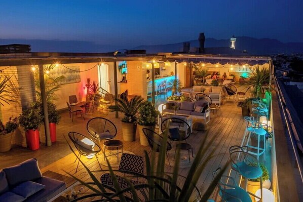 A rooftop bar with outdoor seating, potted plants, and a view of the cityscape at dusk.