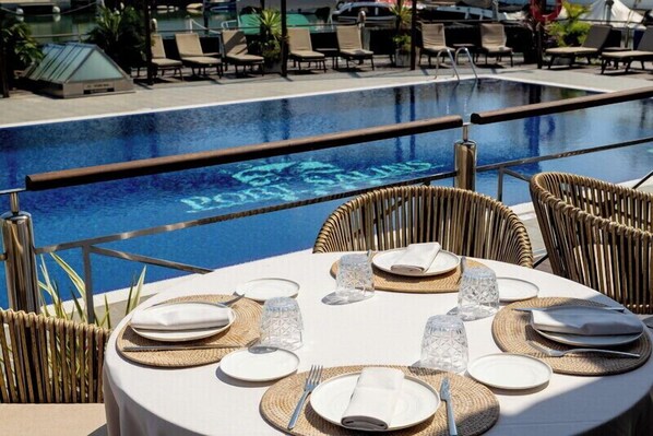 Une table à manger dressée avec des assiettes, des couverts et de la verrerie, donnant sur une piscine ornée du logo « Port of Spain ».