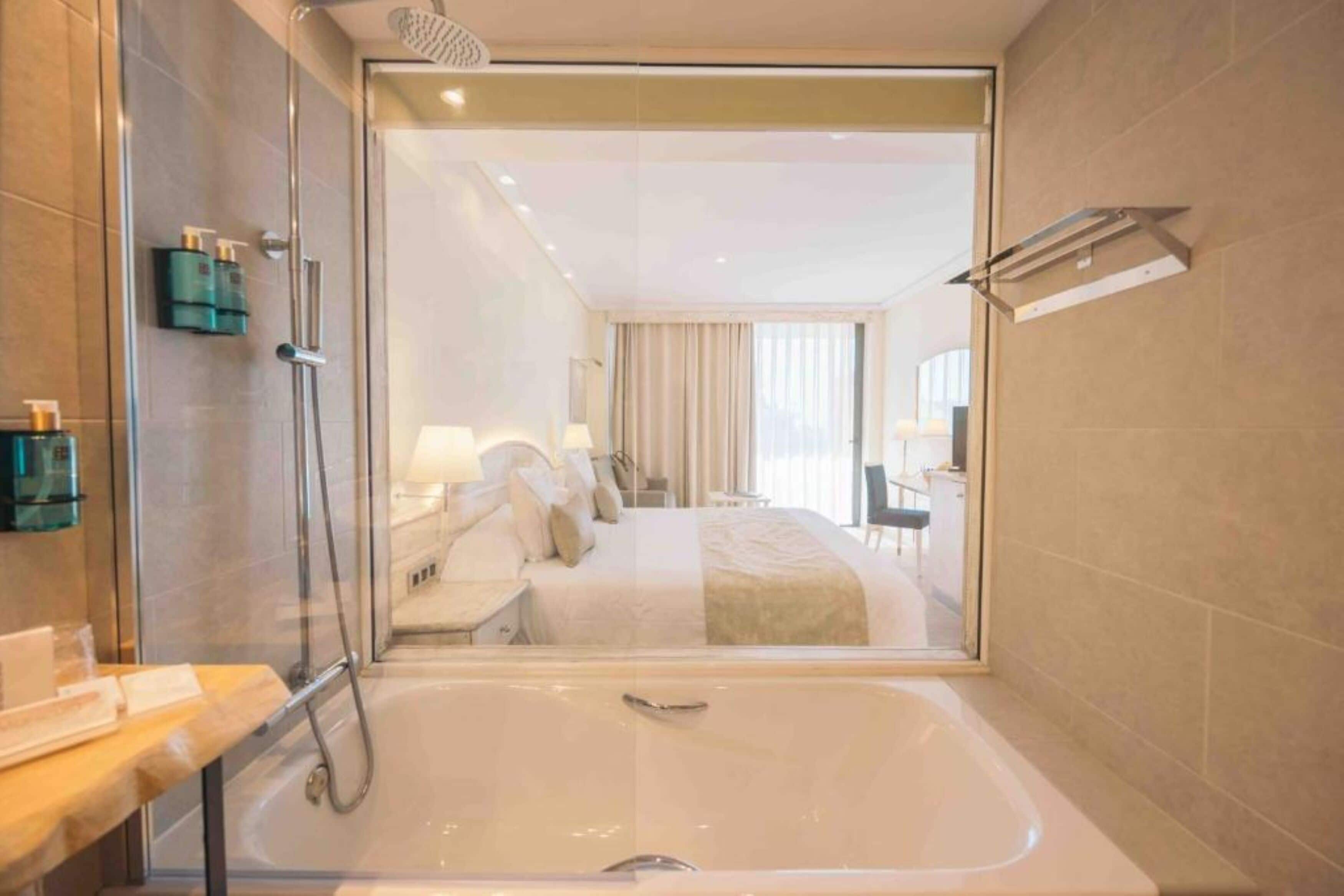 Un baño de hotel moderno con ducha de vidrio, una bañera grande y un dormitorio visible con cama y cortinas.