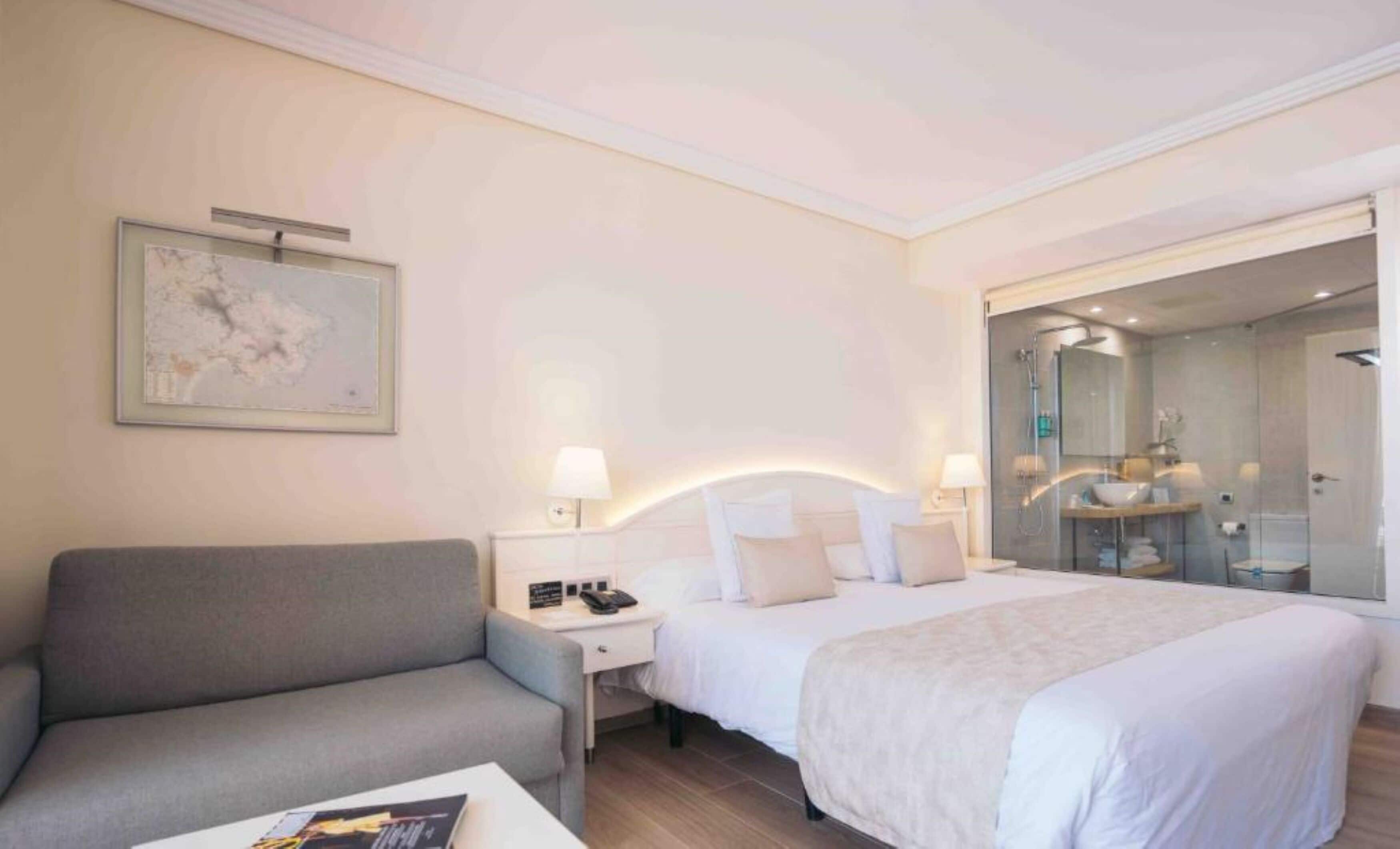 Ein Hotelzimmer mit Bett, einer Couch, einem Nachttisch, einer Lampe und einem durch eine Glastür einsehbaren Badezimmer.