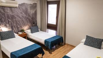 Quarto de hotel com duas camas de solteiro, uma mesinha de cabeceira de madeira, ar condicionado fixado na parede e uma janela com cortinas.