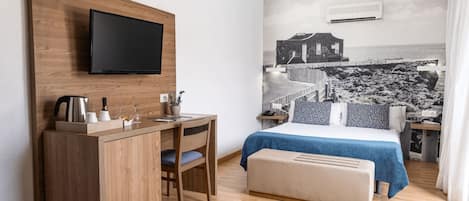 A hotel room with a bed, a desk, a chair, a television mounted on the wall, and a mural of a coastal scene.