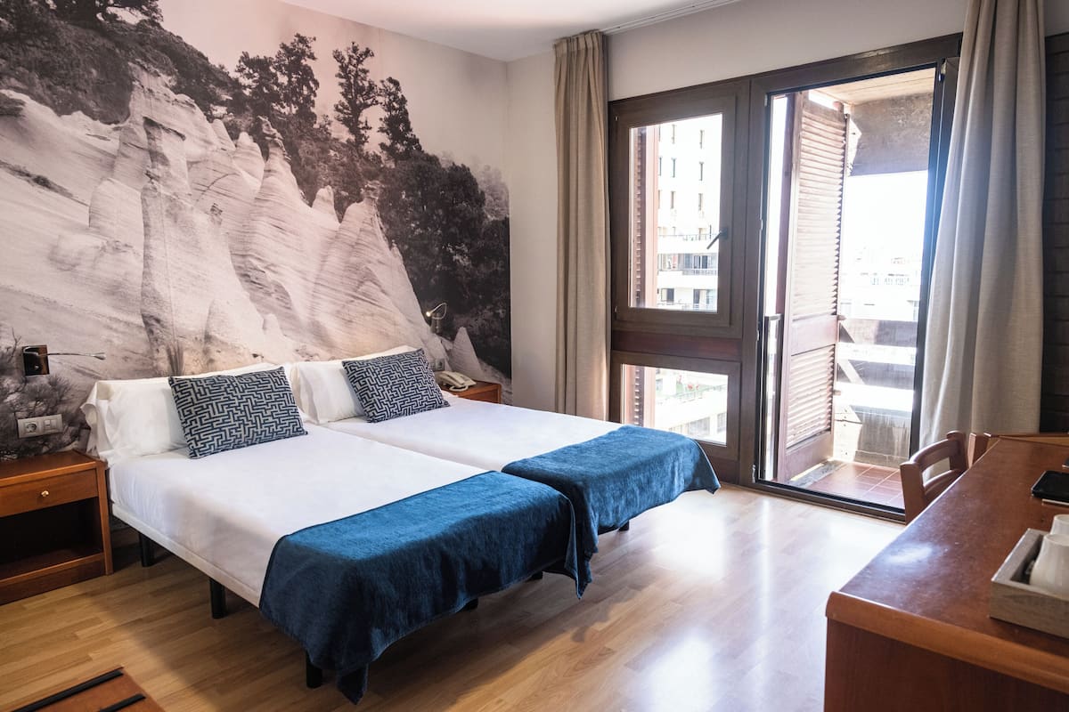 Ein Hotelzimmer mit zwei Betten, einem großen Fenster, einem Holztisch und einem Wandgemälde.