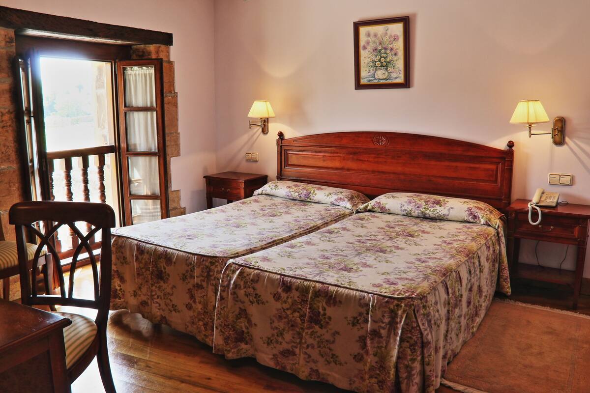 A double bed with floral bedding, two bedside tables with lamps, a wooden chair, and a window with curtains.