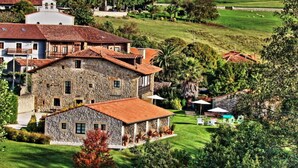 A stone building with a red-tiled roof, surrounded by lush green fields and a few scattered trees. - Palación de Toñanes (Alfoz de Lloredo)