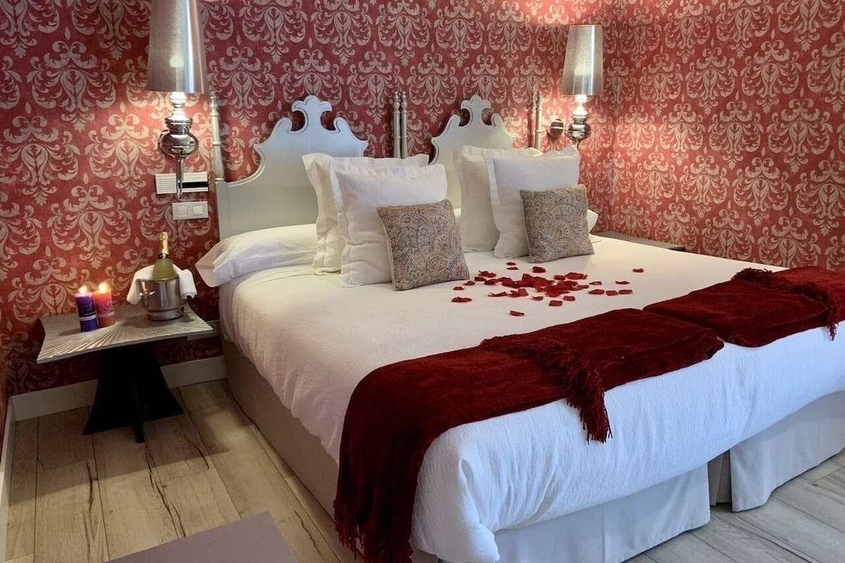 A neatly made bed with white linens and a red throw blanket, a bedside table with a bottle and candles, and a patterned wallpaper background.