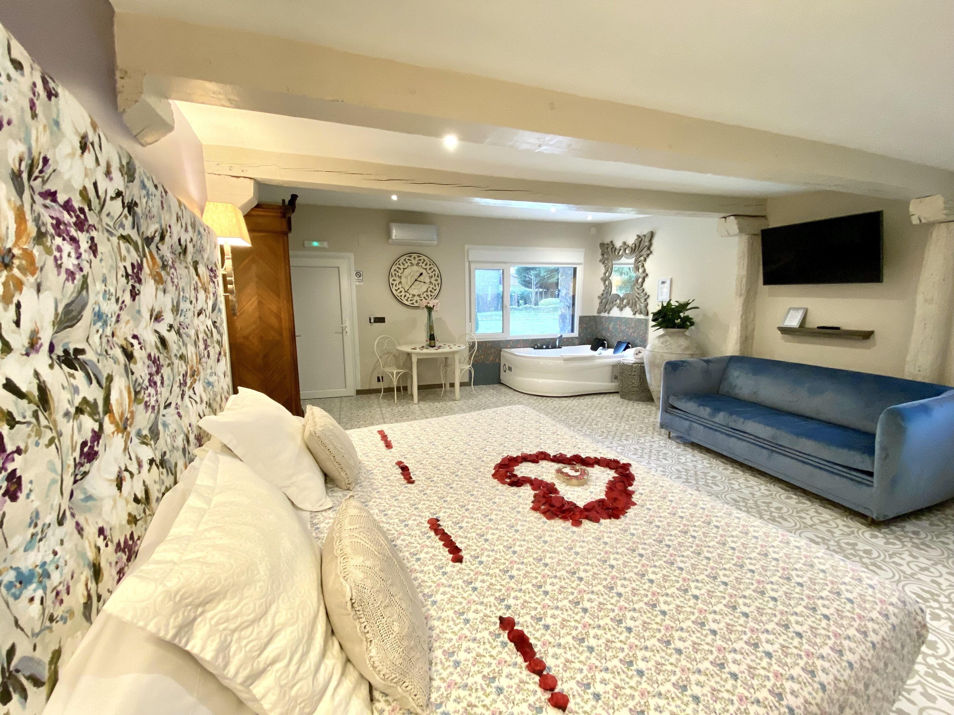A hotel room with a bed, a blue sofa, a heart-shaped decoration on the floor, a dining area with a table and chairs, and a large window.