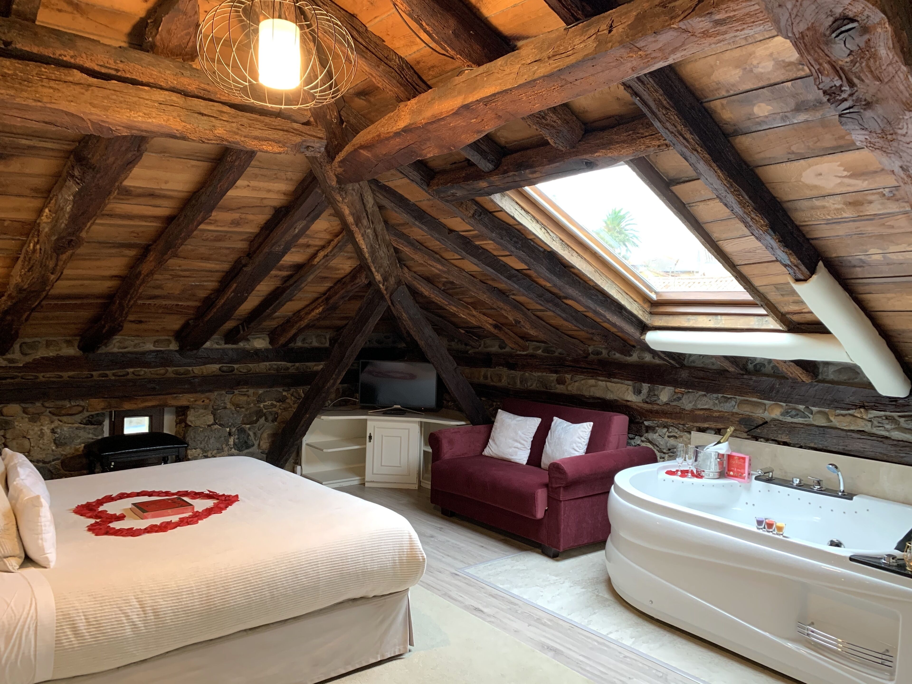 Quarto rústico com parede de pedra, cama com decoração de coração vermelho, sofá vermelho, banheira branca e claraboia.
