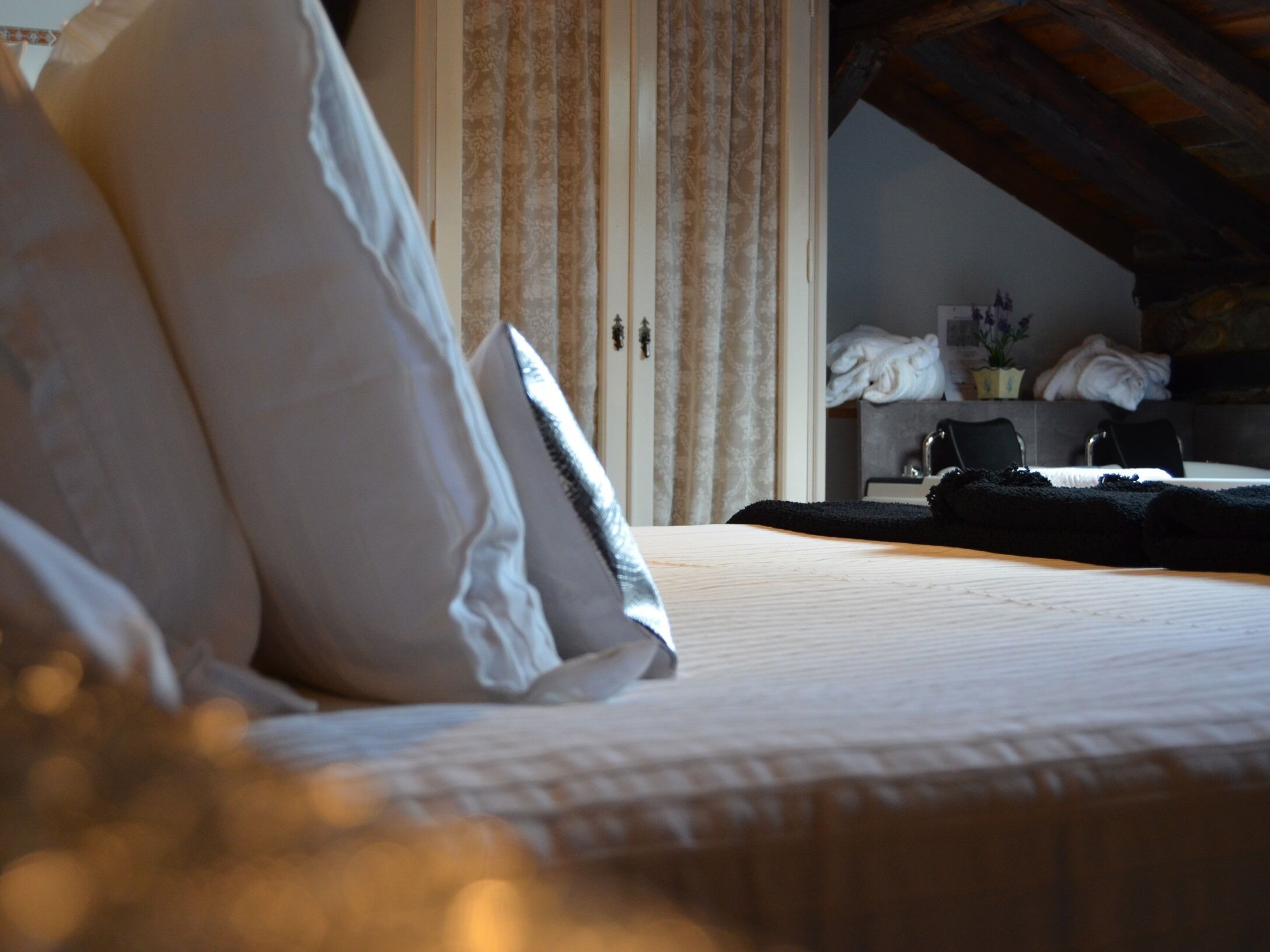 A bedroom with a bed, pillows, a wooden ceiling, and a view of a bathroom with towels and a chair.