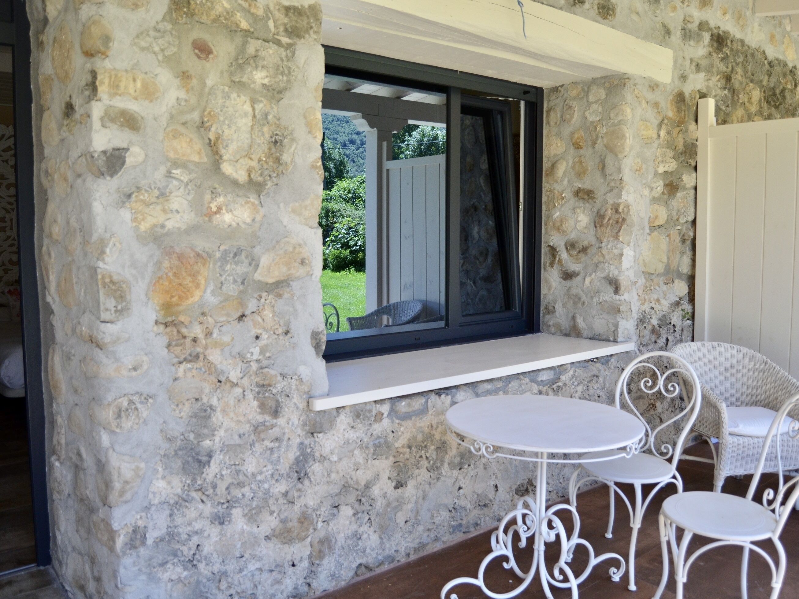 Pared de piedra con una ventana y un espacio exterior con asientos blancos, sillas de metal y una mesa.