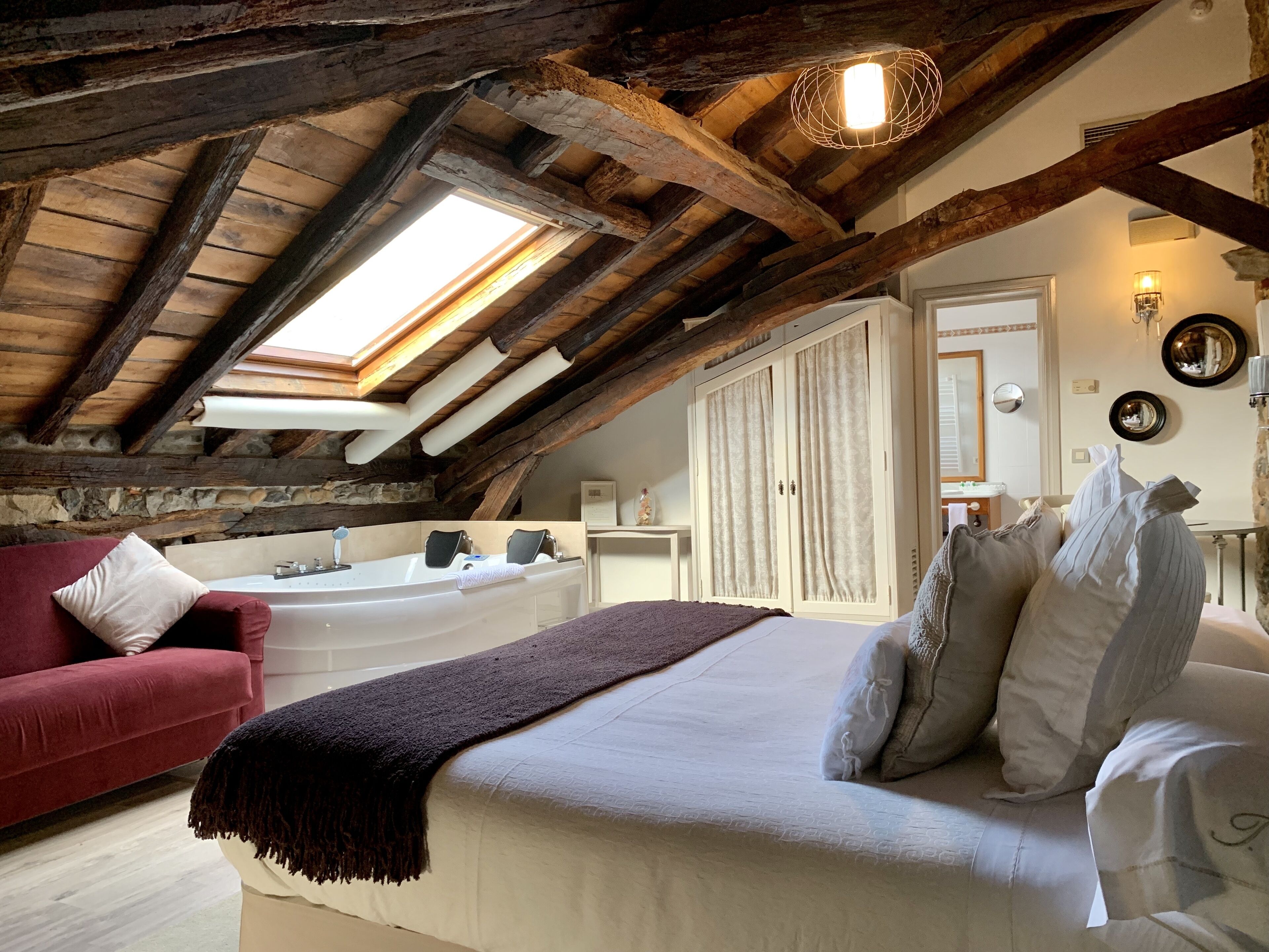 A bedroom with a sloped ceiling, a large window, a bed, a red armchair, a bathtub, and a wardrobe.